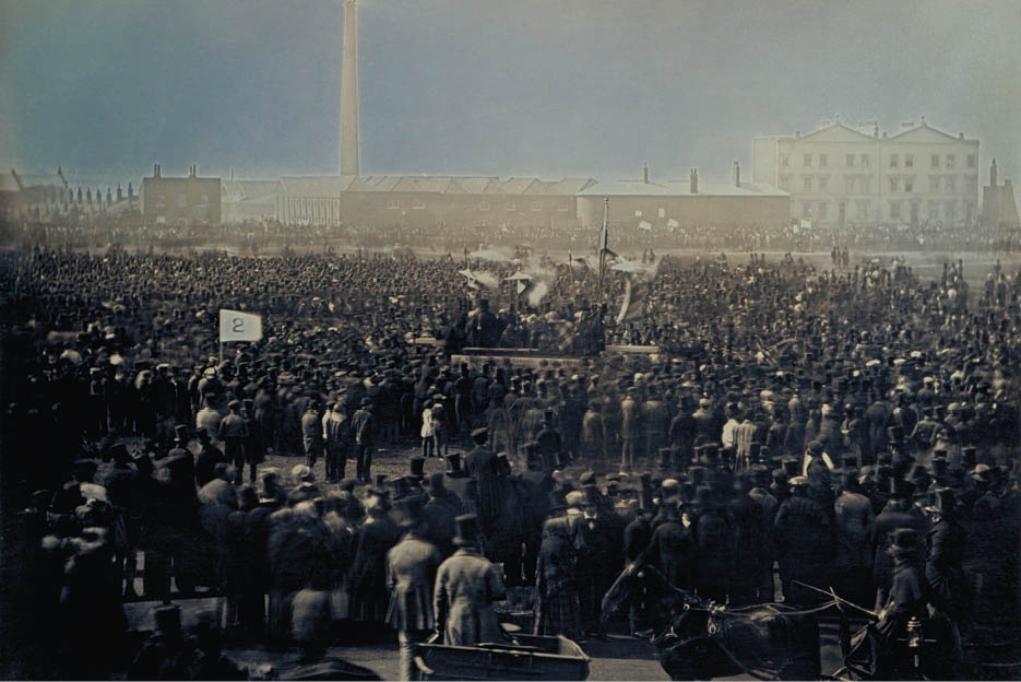 A large crowd of people dressed in early 20th-century clothing gathers outdoors around a platform with speakers and flags in a historic industrial town setting. Description generated by AI