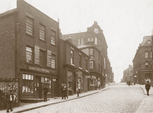 A historic cobblestone street lined with brick buildings and shops features several people walking and standing near the storefronts, with a large clock tower visible in the background. Description generated by AI