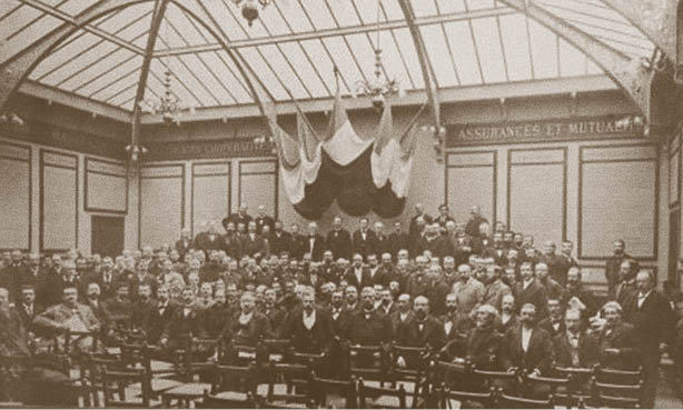 A large group of people dressed in formal early 20th-century attire are seated and standing in a spacious room with a glass ceiling and draped fabric hanging from the center. Description generated by AI