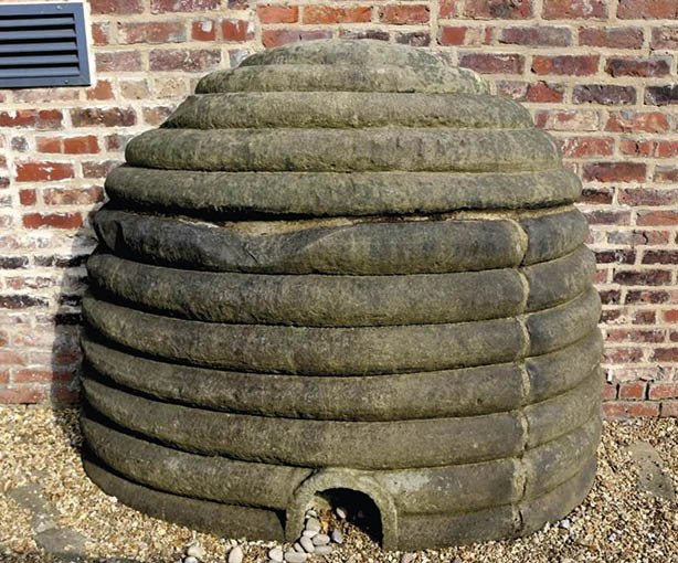 A large stone sculpture resembling a beehive with horizontal ridges and a small arched entrance at the bottom is set against a brick wall on a gravel surface. Description generated by AI