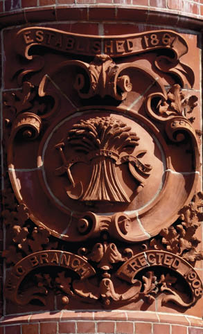 A decorative brick plaque features a carved sheaf of wheat with ribbons and the inscriptions “Established 1867" and "No Branch Erected 1908." Description generated by AI