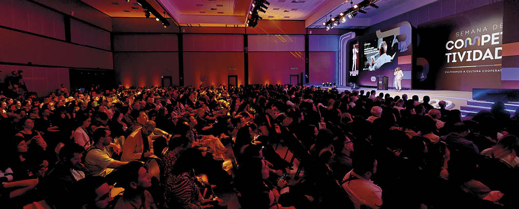 Audit rio cheio com muitas pessoas sentadas assistindo a uma apresenta o em um palco iluminado com proje  es visuais ao fundo.  Description generated by AI