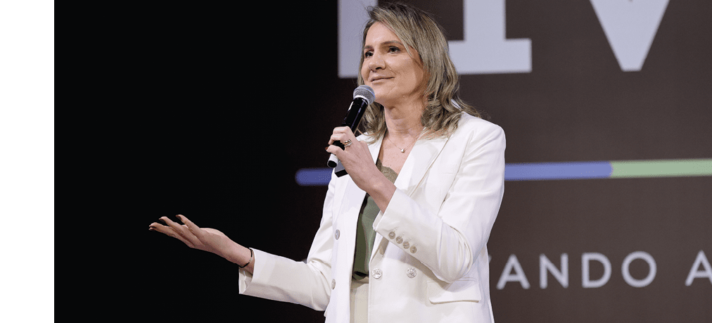 Pessoa adulta com cabelo m dio e ondulado usa terno branco e segura microfone enquanto fala em um evento com fundo escuro e texto parcialmente vis vel. Description generated by AI