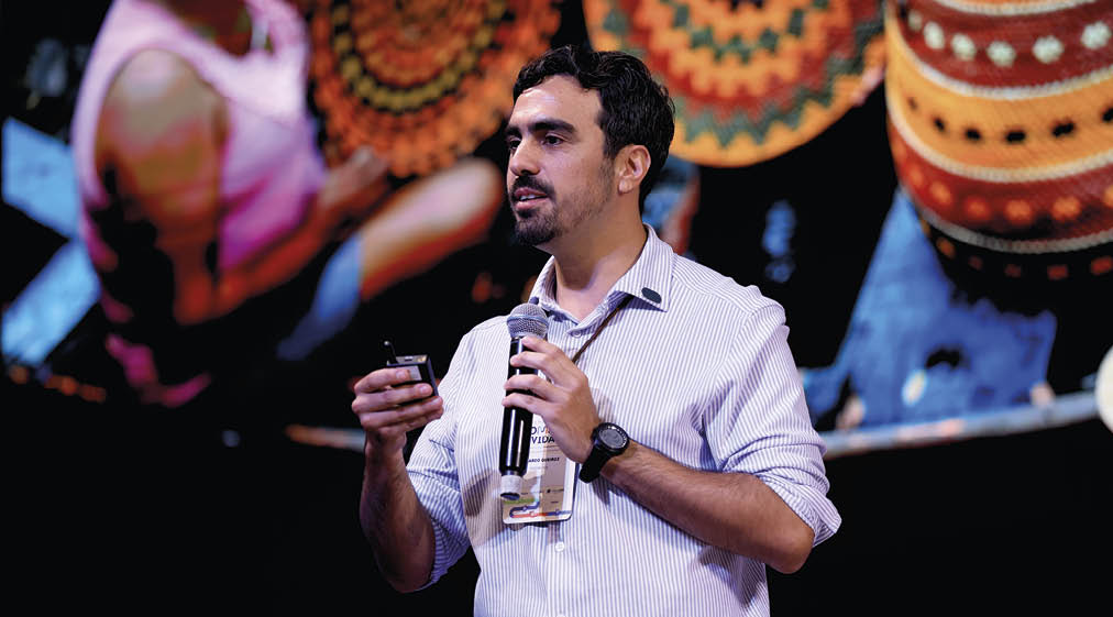 Pessoa adulta com cabelo curto e barba curta segura microfone e controle remoto enquanto fala em um palco com fundo colorido. Description generated by AI