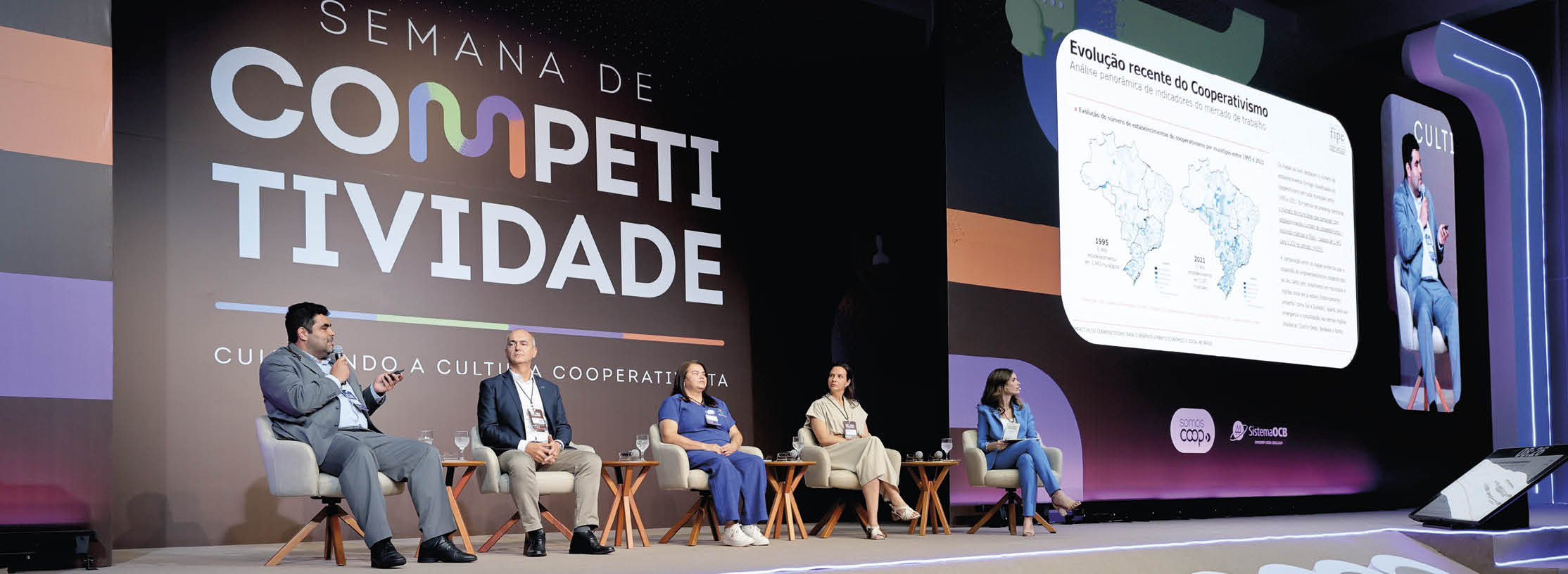 Cinco pessoas sentadas em cadeiras em um palco participam de um painel durante o evento Semana de Competitividade com um tel o exibindo informa es ao fundo.  Description generated by AI