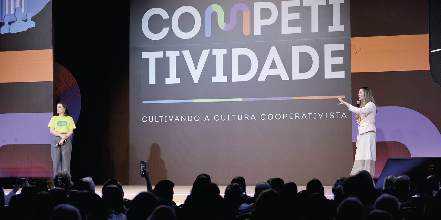 Duas pessoas est o no palco de um evento com um grande painel ao fundo exibindo a palavra “COMPETITIVIDADE" e a frase "CULTIVANDO A CULTURA COOPERATIVISTA" enquanto uma delas fala para a plateia. Description generated by AI