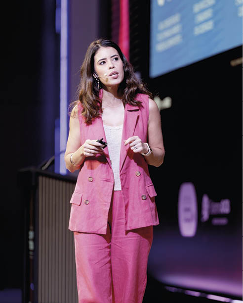 Pessoa adulta com cabelo longo solto vestindo conjunto rosa e camiseta branca falando em evento com microfone de cabe a e segurando controle remoto. Description generated by AI