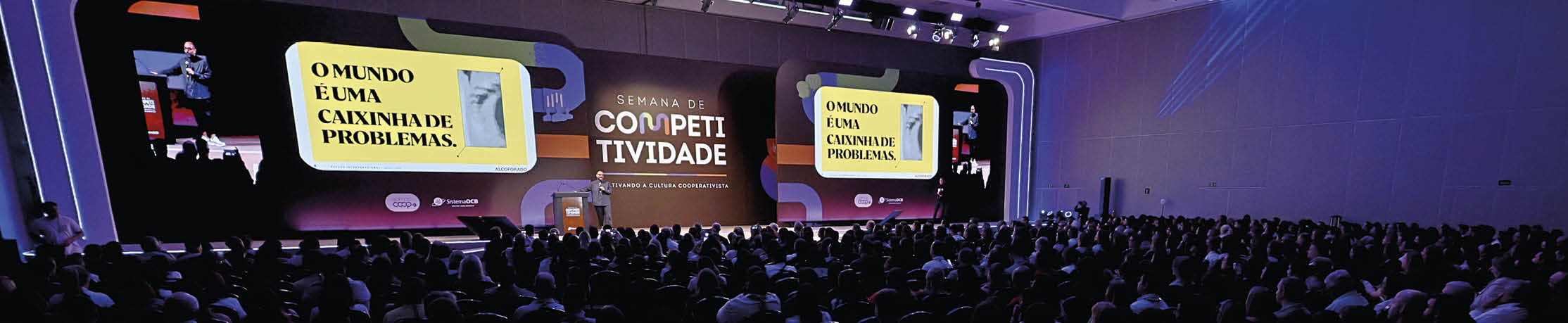 Audit rio cheio com pessoas assistindo a uma apresenta o sobre competitividade exibida em duas grandes telas com texto em portugu s.  Description generated by AI