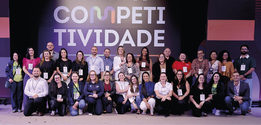 Grupo de adultos reunidos em palco sorrindo para a foto com um fundo escuro e a palavra “COMPETITIVIDADE" em letras grandes iluminadas atr s deles. Description generated by AI