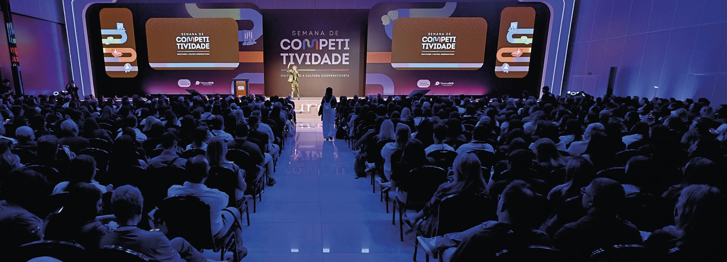 Audit rio cheio com pessoas sentadas assistindo a uma apresenta o em palco sobre competitividade, com telas grandes exibindo informa  es ao fundo.  Description generated by AI