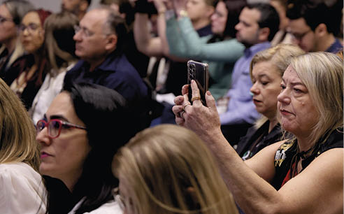 Pessoas sentadas em um ambiente interno parecem atentas, com uma delas segurando um celular para registrar ou fotografar algo  frente.  Description generated by AI