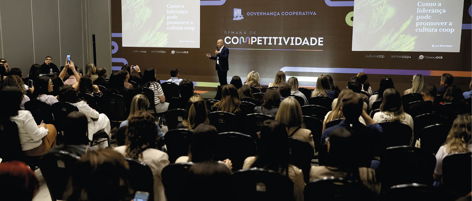 Pessoas sentadas assistem a uma palestra sobre governan a cooperativa e competitividade em um audit rio com um palestrante em p ao centro do palco.  Description generated by AI