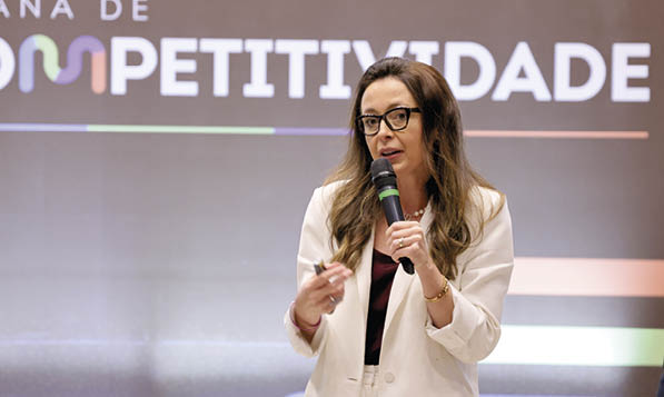 Pessoa com cabelo longo e culos fala ao microfone durante apresenta  o em evento sobre competitividade.  Description generated by AI