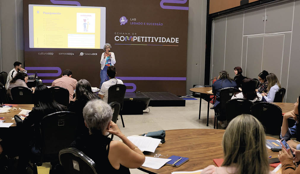 Adultos sentados em mesas redondas assistem a uma apresenta o em um audit rio com um tel o que exibe um slide sobre competitividade.  Description generated by AI
