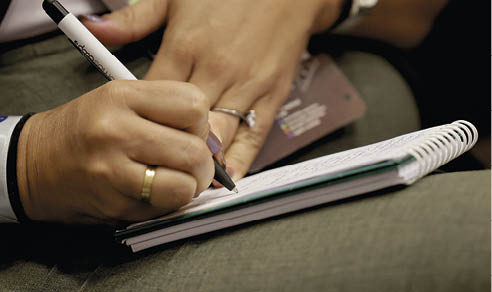 Pessoa escrevendo em caderno espiral com caneta preta enquanto segura um celular. Description generated by AI