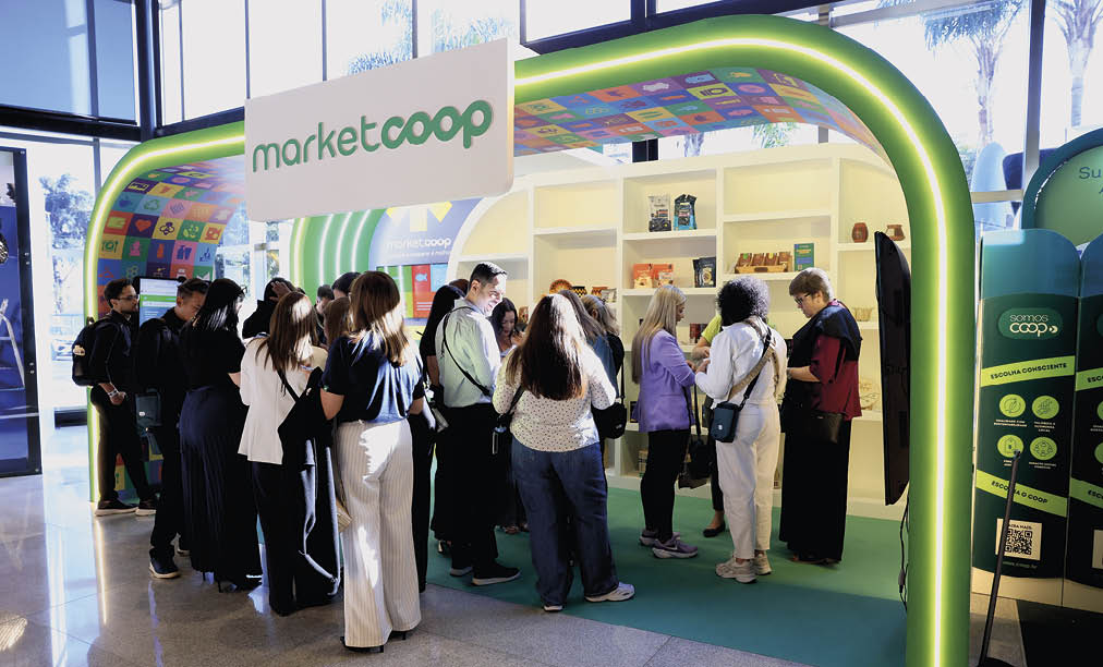 Pessoas em p conversando e interagindo em um estande colorido com a marca marketcoop em um ambiente interno iluminado por luz natural.  Description generated by AI
