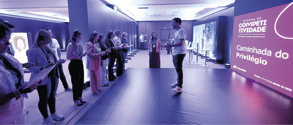 Pessoas em p em fila segurando cadernos enquanto uma pessoa fala em um palco em ambiente interno com ilumina  o azul e um painel digital ao fundo com texto sobre caminhada do privil gio.  Description generated by AI