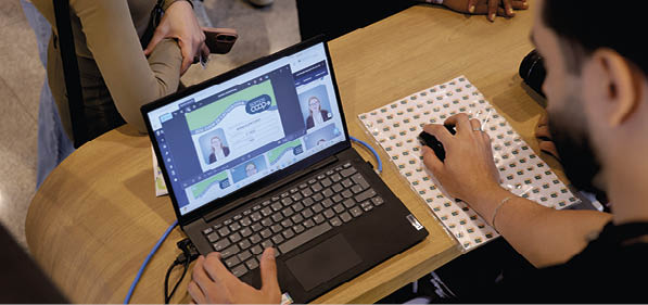 Pessoa usando laptop em uma mesa de madeira enquanto participa de uma videoconfer ncia, com outras pessoas ao redor. Description generated by AI