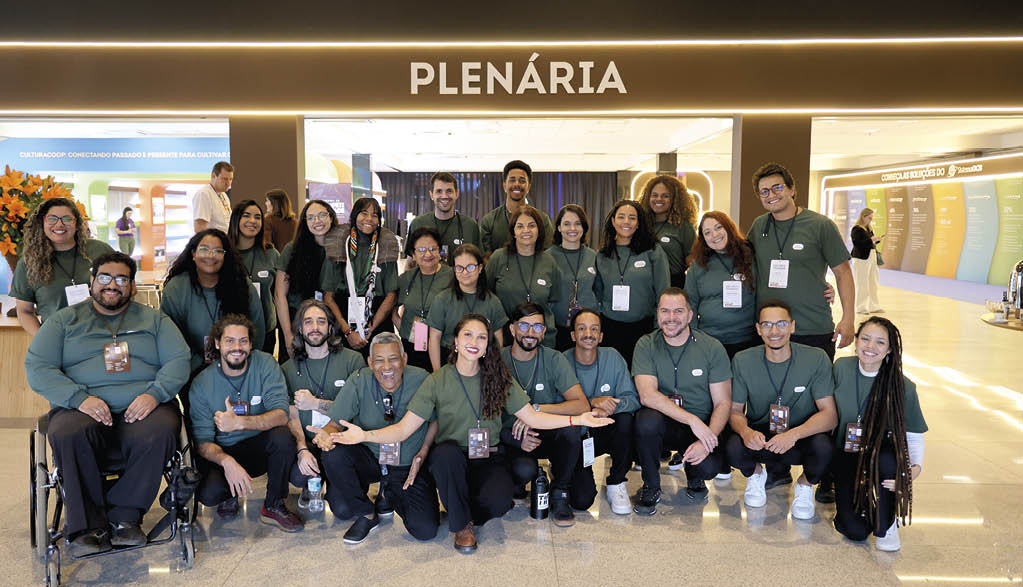 Grupo de pessoas sorridentes usando camisetas verdes posando para foto em ambiente interno com placa iluminada escrita Plen ria ao fundo. Description generated by AI