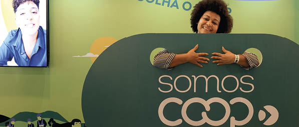 Pessoa com cabelo cacheado sorri enquanto aparece atr s de um painel verde com a frase somos coop em um ambiente com fundo verde e azul com texto relacionado a escolha consciente. Description generated by AI