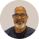 Pessoa adulta com cabelo curto grisalho, barba branca, culos de arma  o preta e camiseta azul escura, sorrindo levemente com fundo circular bege.  Description generated by AI