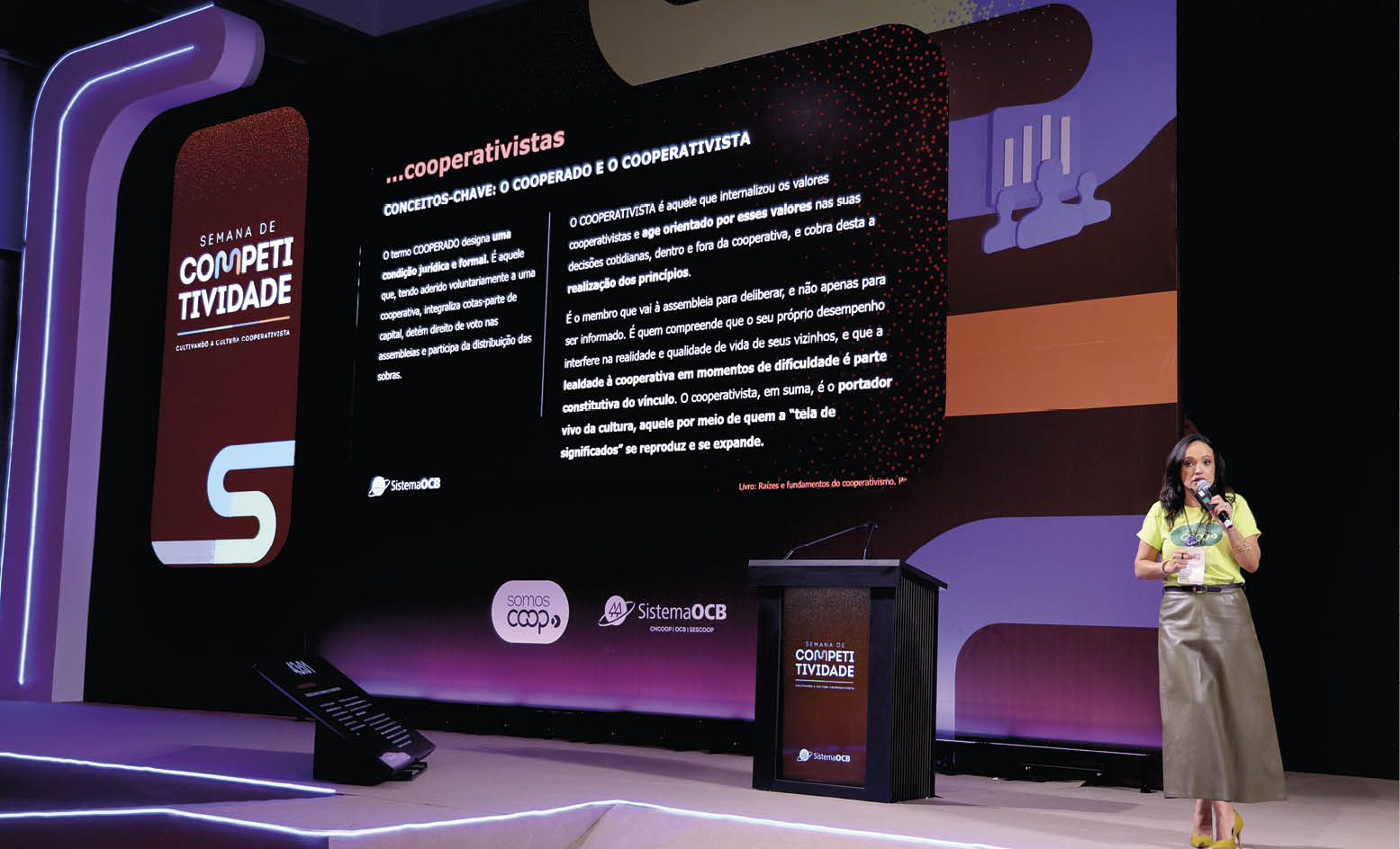Pessoa com camiseta amarela e saia longa bege fala em microfone em palco iluminado com painel digital exibindo texto sobre cooperativismo. Description generated by AI