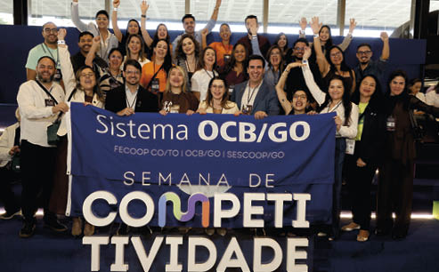 Grupo de adultos sorridentes posando em ambiente interno com banner azul que diz Sistema OCB/GO e Semana de Competitividade. Description generated by AI