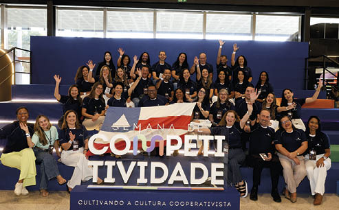 Grupo de adultos sorridentes sentados e em p em degraus coloridos segurando uma faixa com o texto “Semana de Competitividade" em um ambiente interno iluminado.  Description generated by AI