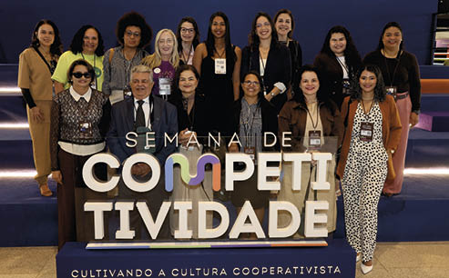 Grupo de pessoas sorrindo posando em um evento chamado Semana de Competitividade com um letreiro grande e colorido ao centro e um fundo azul escuro. Description generated by AI