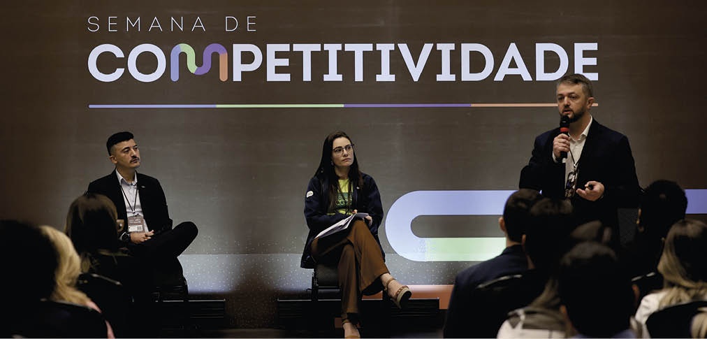Tr s adultos participam de um painel de discuss o durante a Semana de Competitividade, com um deles falando ao microfone enquanto os outros dois escutam sentados. Description generated by AI