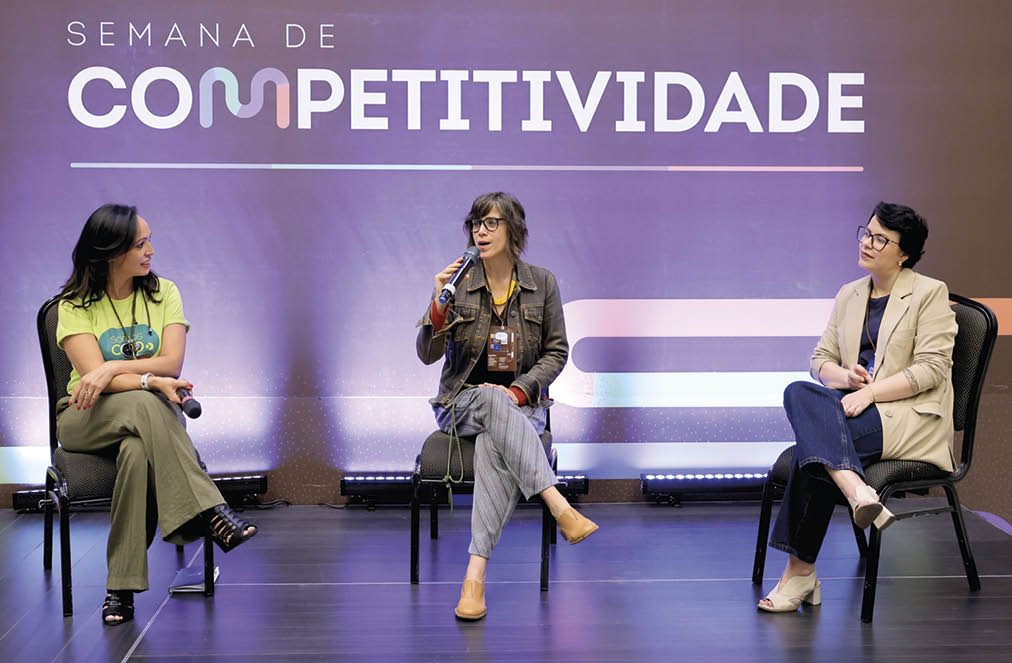 Tr s adultos sentados em cadeiras em um palco participam de um painel durante a Semana de Competitividade, com um fundo roxo e ilumina o suave.  Description generated by AI
